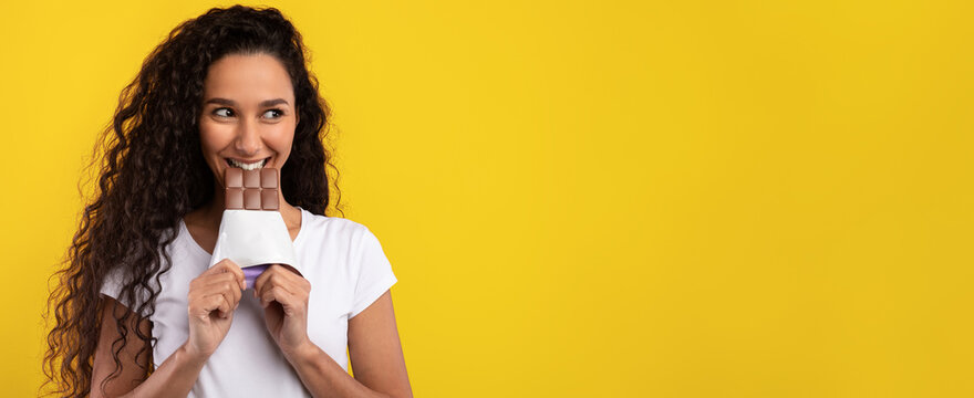 A woman with curly hair smiles while playfully holding a chocolate bar in front of her mouth. The bright yellow background adds a fun and cheerful atmosphere to the scene.