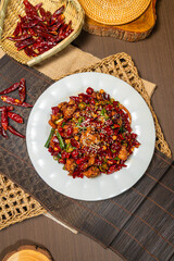 A plate of spicy stir-fried chicken garnished with sesame seeds, surrounded by dried red and green chilies, served on a rustic woven mat.