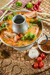 Thai-style Tom Yum soup in a traditional hot pot with shrimp, lemongrass, chili, garlic, lime, and herbs, served on ornate Thai fabric.
