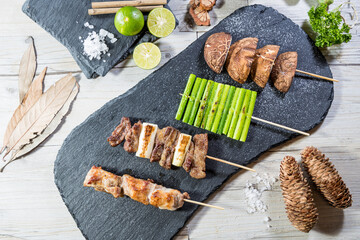 A slate board with three skewers: grilled meat with leeks, asparagus, and mushrooms, surrounded by lime, salt, bay leaves, and cinnamon sticks.