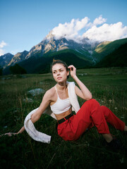 Fototapeta premium Meadow scene with mountain backdrop, a woman sits on grass in a white top and red pants, a towel draped over her shoulder, serene outdoor mood under a blue sky