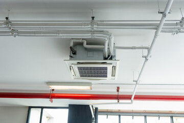 Ceiling Cassette Air Conditioner with Exposed Ductwork and Utility Pipes, HVAC and Fire Safety System Integration on Modern White Commercial Ceiling