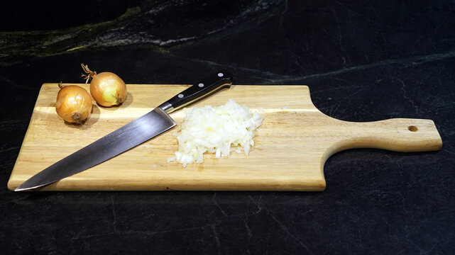 Two whole, peeled onions and a pile of freshly diced onion rest next to a chef's knife on a wooden cutting board against a dark countertop