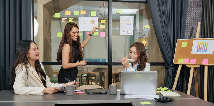 Business women meet, busy team people work on project, presenting data with colorful charts and sticky notes on glass wall, discussing strategy in bright modern conference room