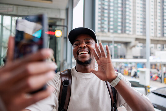 Smiling man waves during video call in urban setting.