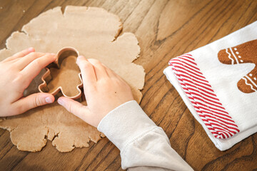Child’s hands using a cookie cutter to shape gingerbread dough on a wooden table during Christmas baking.