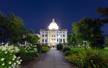 Illuminated South Dakota State Capitol building in Pierre at dusk. Walkway with flowers leads to the building, framed by trees under a clear blue sky