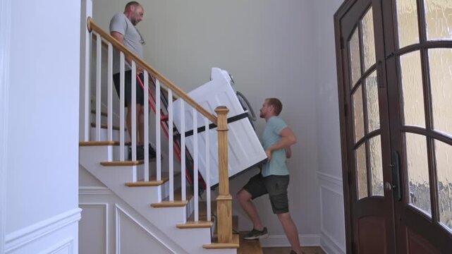 team carefully relocates washer outside, residents coordinate to carry appliance safely outside, daylight illuminates workers as they carefully transport appliance past banister and doorway