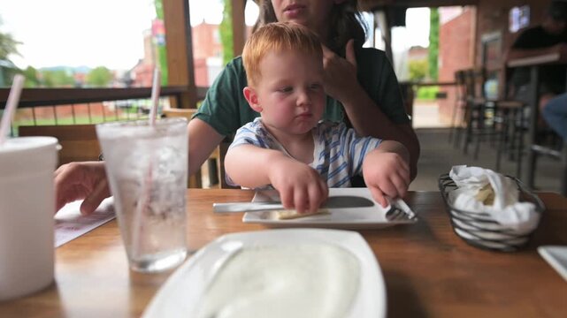 caucasian mother and toddler waiting meal at outdoor restaurant, child fidgets with napkin and utensils while mother arranges plate, wooden table and daylight set scene of anticipation