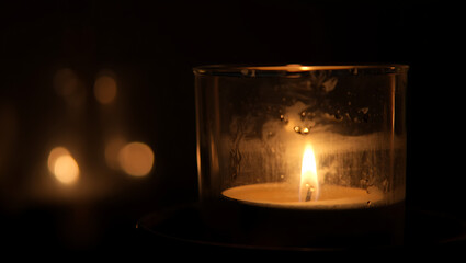 A single warm glowing candle flame illuminates a dark room casting soft light and creating beautiful bokeh reflections on the left side of the frame