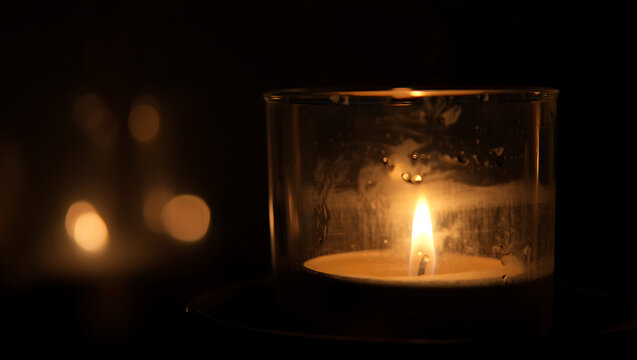 A single warm glowing candle flame illuminates a dark room casting soft light and creating beautiful bokeh reflections on the left side of the frame