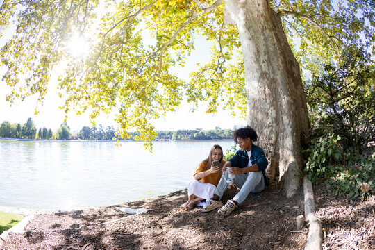 Couple of friends sitting by a lake hanging out using smartphone