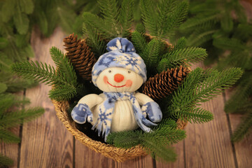 A soft snowman toy with a blue hat and a snowflake-patterned scarf lies in a wicker basket among fir branches and pine cones on a wooden background.