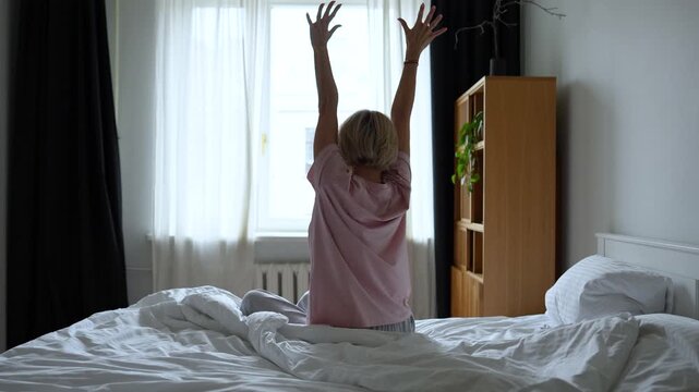 Rested middle aged woman with smiling expression waking up in early morning, sitting in bed stretching arms and body after good sleep, looking at window, getting ready to begin day with positivity.
