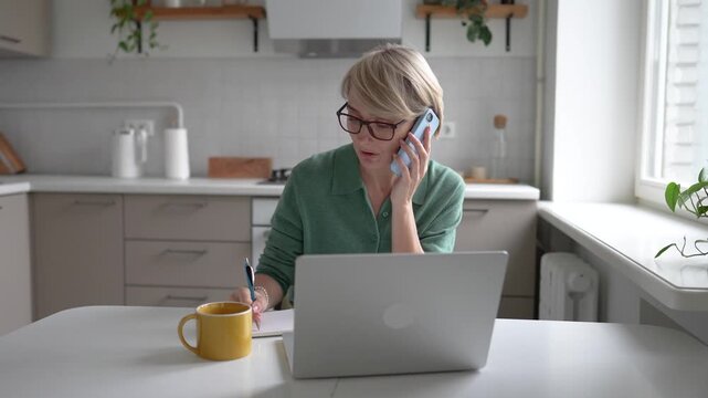 Middle aged businesswoman working multitasking on laptop, writing notes in notebook and gets distracted with smartphone, checking messages, answering call, having troubles with concentration and ADHD.