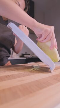 Close-up of Asian woman shredding cabbage using a mandoline slicer on a wooden cutting board in a home kitchen, vertical format