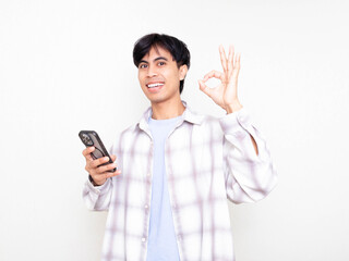 Fototapeta premium Young man holding a smartphone and showing an OK hand gesture while smiling, expressing approval and positive confirmation in a bright, clean setting.