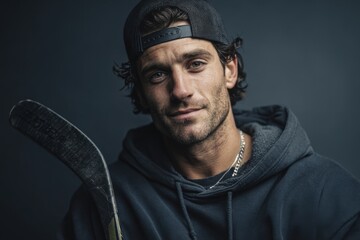 A man wearing a hoodie and a backwards ball cap. He is holding a hockey stick.