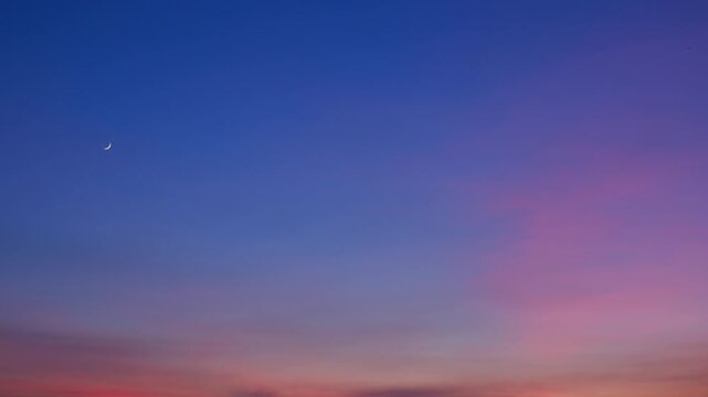 A serene sunset sky timelapse featuring soft cirrus clouds glowing pink and purple, blending into warm horizons with a delicate crescent moon enhancing the tranquil evening atmosphere. Time lapse.
