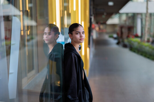 Young woman in black jacket leans against glass wall.