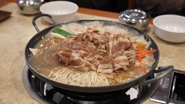 Korean Bulgogi Jeongol Hot Pot with Thinly Sliced Pork, Noodles, and Vegetables, Shabu-shabu Style Meat and Enoki Mushrooms Boiling in Soup Base, Meal Served in Cast Iron Pot on Gas Burner