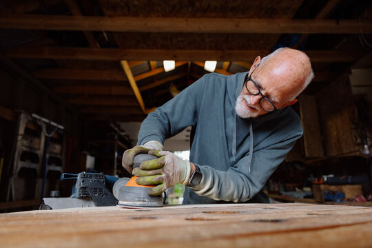 Candid elderly woodworker sanding plank in workshop with belt sander