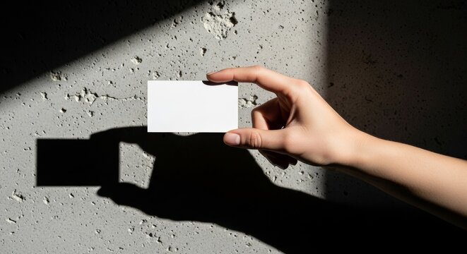 A hand holding a blank white business card against a textured concrete wall with shadows.