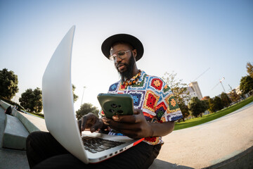 Black man multitasking using laptop and smartphone outdoors