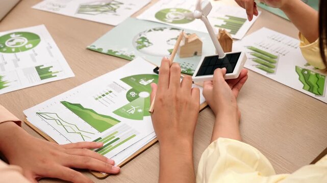 Business team focus windmill model, represented using clean energy. Coworker analyze green power graph choosing way to decrease carbon. Wooden block symbolized ecology house put on table. Delineation.