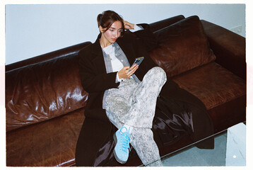 Woman sitting on a brown sofa looking at her phone, autumn outfit 