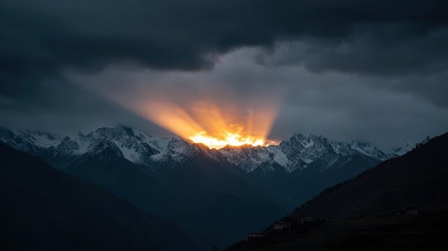 Brilliant sun rays emerge from behind snowcapped mountain peaks under a dark sky