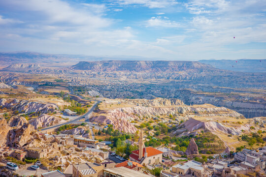 A vibrant panoramic view of Cappadocia, Turkey, showcasing its iconic rock formations, fairy chimneys, rolling valleys, and a charming village with a mosque tower under a bright blue sky