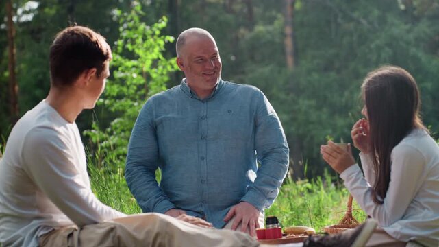 Happy father enjoying outdoor picnic with son and daughter, smiling while discussing warmly, girl sipping tea from cup and boy nodding with cheerful expression under sunlight
