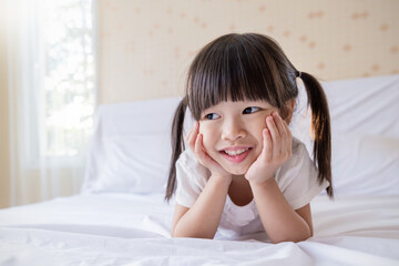 Portrait adorable little girl with pigtails smile brightly while lying on a white bed in a cozy cheerful home environment. Education preschool toddler.