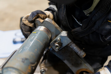 Mechanic using gloved hands to service hydraulic cylinder joint, focusing on metal threads, hoses, and industrial fittings during essential maintenance procedures on heavy equipment machinery