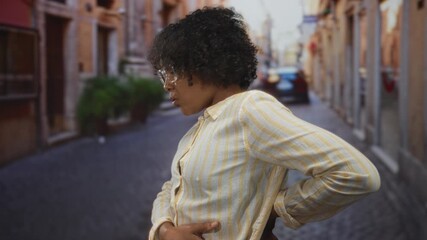 Young african american woman holds lower back in pain gesture on a narrow cobblestone street in sunlit city lane; pain discomfort.