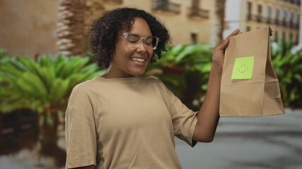 Woman wearing glasses holds paper bag with smiley note on city street; happiness generosity kindness.