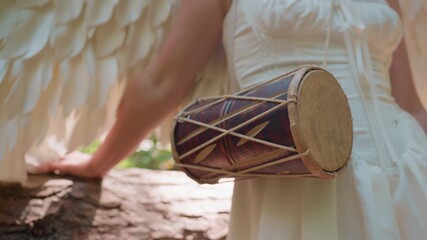 Close up of lady with angel wings carrying traditional talking drum while resting hand on fallen tree trunk in warm forest sunlight, representing peace, spirituality and unity with natural world