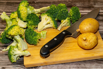 Fresh broccoli florets and whole potatoes sit on a wooden cutting board next to a chef's knife, ready to be prepared for a healthy meal.