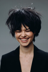 Smiling woman with short dark hair laughing joyfully against gray background in black outfit.