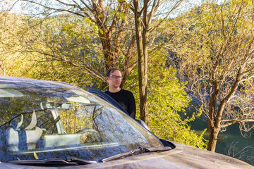 A traveler stands by a parked car in a forested area, enjoying a quiet outdoor moment during a road and hiking trip.