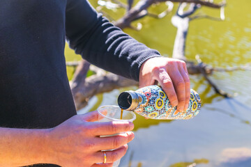 Hands pouring drink from patterned thermos into plastic cup near water during outdoor rest.