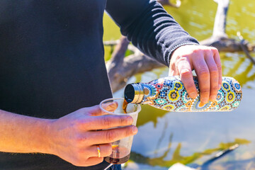 Hands pouring drink from patterned thermos into plastic cup near water during outdoor rest.