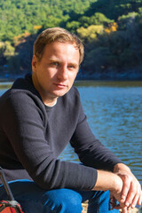 Person sitting by lake in nature, resting near water with forest on background.