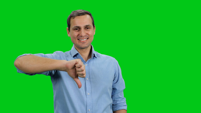 Young man in light-colored shirt smiling condescendingly but shows his disagreement with thumbs down gesture sign on green background