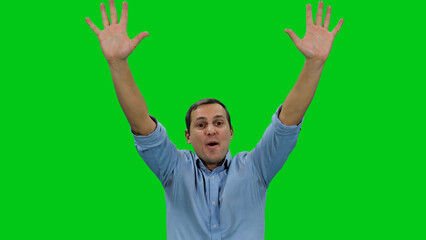 Happy emotional young adult man in light blue colored shirt  raised hands with joy against green background