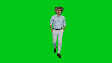 Young adult smiling man tourist walking in light-colored shirt, pants and cowboy hat with hands in pockets against green background