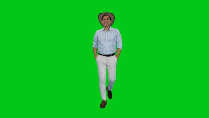 Young adult smiling man tourist walking in light-colored shirt, pants and cowboy hat with hands in pockets against green background