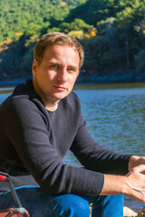 Person sitting by lake in nature, resting near water with forest on background.