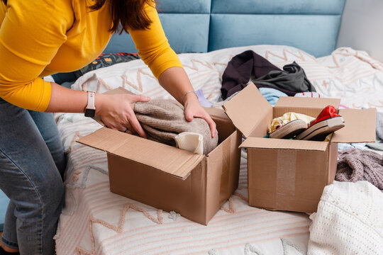 A young woman sorting clothes and packing into cardboard boxes. Donations for charity, help low income families, declutter home, sell online, moving moving into new home or recycling